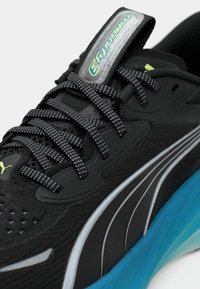 Gros plan sur une basket Puma Magnify 3 noire avec des lacets réfléchissants, des rayures grises et un détail de semelle bleu vif.