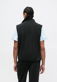 Vest de fleece negru cu guler înalt și laturi deschise, purtat peste o cămașă cu mâneci scurte de culoare bleu deschis, având o textură netedă și branding minimal.