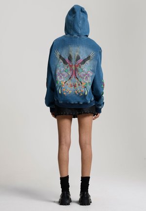 Person wearing blue denim hoodie with colorful bird and floral embroidery on back, paired with black skirt, black socks, and chunky shoes.