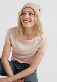 Lichtroze t-shirt met korte mouwen van zachte stof, gecombineerd met een geribde lichtroze beanie. Het model heeft golvend haar en zit met gekruiste benen.