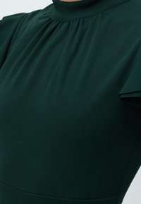 Robe vert foncé à col montant avec manches courtes et légères fronces sous l’encolure, dévoilant le haut de la poitrine et la zone des épaules.