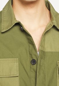 Camicia verde oliva con colletto a bottoni, con bottoni neri e tasche sul petto. Il tessuto appare texture e resistente.