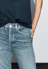 Jean en denim slim fit, bleu clair, taille haute, avec cinq poches et des détails de couture subtils. Le matériau a une finition lisse.