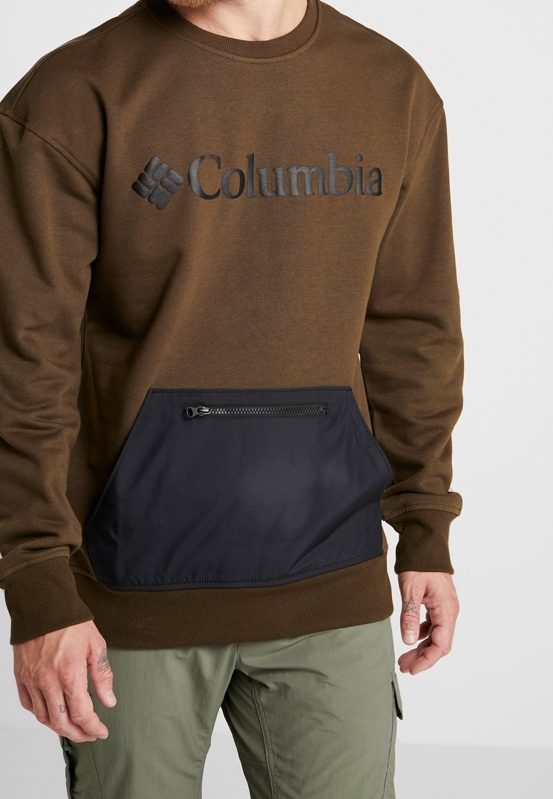 Homme portant un sweat-shirt Columbia marron avec une poche avant zippée noire et un pantalon cargo vert olive, recadré aux épaules et aux genoux.
