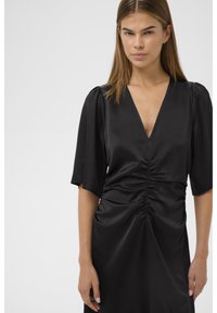 Musta mekko, jossa syvä V-kaula-aukko ja lyhyet pussihihaiset hihat. Vyötäröllä on laskostettu yksityiskohta ja sileä pinta.