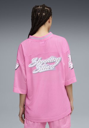 Persoon met gevlochten haar, gekleed in een losse roze mesh sporttrui en bijpassende korte broek, met de tekst "Shooting Starz" op de achterkant.