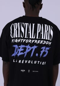 Černé bavlněné tričko s výrazným bílým a modrým textem. Grafika zahrnuje "CRYSTAL PARIS", "DEPT. 75" a "LA RÉVOLUTION" ve worn stylu.
