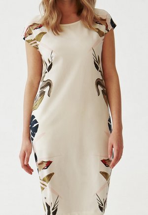 Femme portant une robe couleur crème avec des manches courtes, présentant des motifs floraux abstraits en noir, vert, rouge et marron.