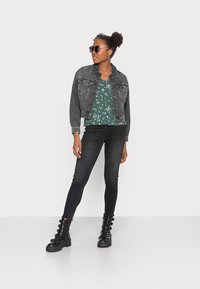Jaqueta de denim preta, blusa verde floral, jeans skinny pretos e botas pretas chunky com detalhes em fivela. Óculos de sol completam o look.