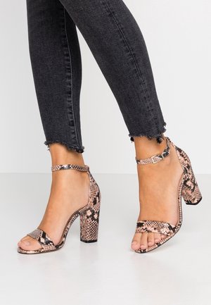Pieds portant des sandales à talons hauts avec un motif peau de serpent rose et noir et des brides à la cheville, associées à un jean noir à ourlet effiloché.