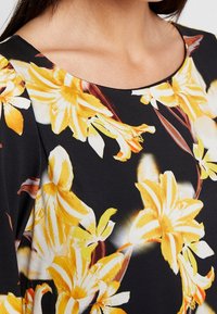 Blusa floral con fondo negro y vibrantes flores amarillas y blancas. Tela suave y lisa con escote redondo y un ligero detalle de fruncido.