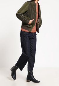 Olivově zelená bomber bunda přes svetr v rezavé barvě, doplněná tmavými denimovými kalhotami a černými kotníkovými botami s metalickým podpatkem.