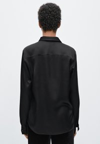 Black satin long-sleeve shirt with a straight cut, point collar, and buttoned cuffs. Smooth texture and rounded hemline at the back.