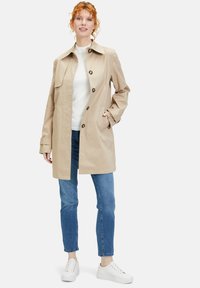 Beige Trenchcoat mit Knopfverschluss, großem Kragen und zwei Seitentaschen. Kombiniert mit blauen Jeans und weißen Sneakers. Glatte Textur.