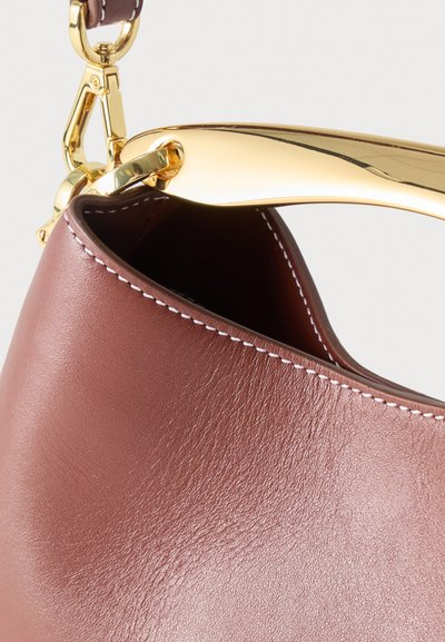 Καφέ δερμάτινη τσάντα χειρός με λευκή ραφή, που διαθέτει λαμπερό χρυσό μεταλλικό χερούλι και κούμπωμα, σε φωτεινό φόντο.