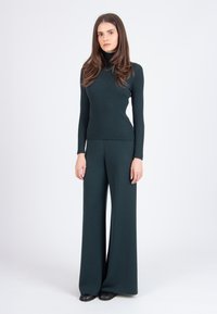 Waldgrüner gerippter Rollkragenpullover kombiniert mit weiter geschnittenen Hosen. Beides ist aus einem dicken Stoff gefertigt und weist ein nahtloses Design auf.