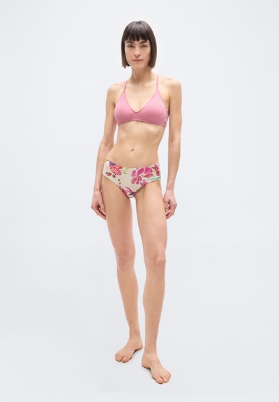 Femme debout pieds nus portant un haut de bikini rose et un bas de bikini taille haute à motifs floraux sur fond blanc uni.