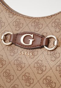 Borsa a tracolla marrone con motivo, caratterizzata da una texture liscia, con un logo G in oro e dettagli in metallo. Nome del marchio "GUESS" impresso sotto.