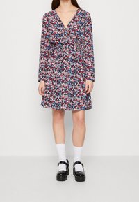 Blommig omslagsklänning med långa ärmar, med ett flerfärgat mönster på en marinblå bakgrund, cinchad midja och A-linjeformad kjol. Svarta plattformsskor bärs.