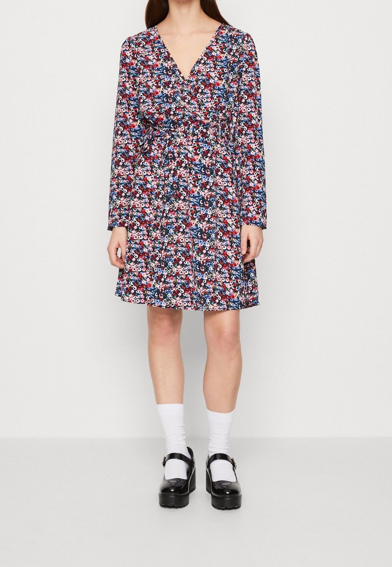 Blommig omslagsklänning med långa ärmar, med ett flerfärgat mönster på en marinblå bakgrund, cinchad midja och A-linjeformad kjol. Svarta plattformsskor bärs.