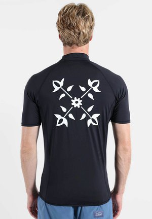 Homme aux cheveux blonds courts portant un t-shirt noir à manches courtes avec un motif croisé floral blanc dans le dos, debout devant un fond uni.