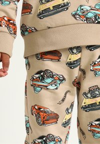 Beige tøj med farverige tegneserie biler i rød, gul og blå. Stoffet har en glat tekstur med ribbede mansjetter.