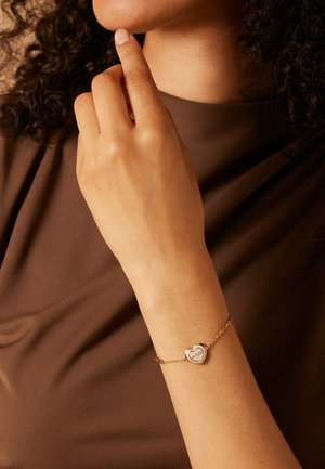 Bracelet en chaîne dorée avec un pendentif en forme de cœur, présentant une surface texturée et des détails gravés, porté sur une main sur un fond marron.