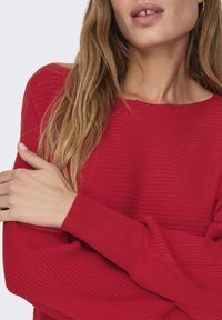 Red ribbed sweater with long sleeves, featuring a wide neckline and textured fabric. Arms are crossed, showcasing the sleeve detail.