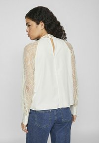 Blusa suelta de crema con mangas largas de encaje transparente, detalle de ojal en la parte posterior y un acabado de tela suave. Combinada con jeans de mezclilla azul.