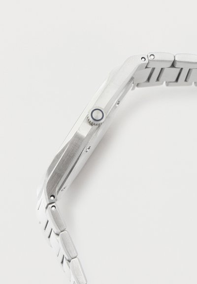 Λεπτό ασημένιο μεταλλικό ρολόι με μπρασελέ από κρίκους και αυλακωτό κορόνα στη μία πλευρά πάνω σε λευκό φόντο.