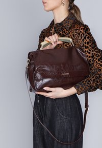 Bruine leren handtas met een glanzende afwerking, handvat in goudkleur, afneembare schouderband en een klep sluiten. De textuur is gerimpeld en glad.