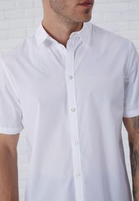 Camisa de botones de manga corta blanca con un cuello clásico y botones transparentes, hecha de una tela suave con un diseño minimalista.