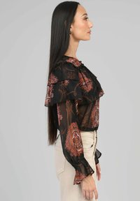 Blouse noire et transparente avec des manches volantes et un motif floral dans des tons de marron et de crème. Détail en nœud au cou, ajustée à la taille.