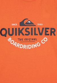 Oranžové bavlněné tričko s černobílým logem „Quiksilver“ a „Boardriding Co“ a rokem „1969“ nad grafickým zobrazením vlny.