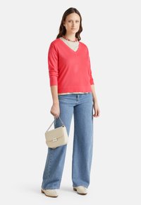 Maglione corallo a scollo a V, tessuto leggero, abbinato a jeans a gamba larga di colore blu chiaro. Porta una borsa crema con dettaglio in catena argentata.