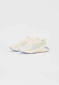 Pár světle béžových sportovních tenisek s bílými tkaničkami, šedými podrážkami a modro-zeleným pruhovaným logem Puma na boku.