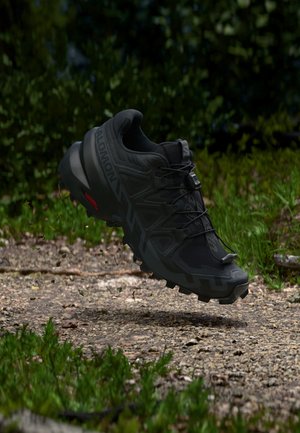 Chaussure de trail running Salomon noire en l'air au-dessus d'un sentier forestier gravillonné avec un feuillage vert en arrière-plan.