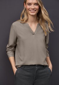 Pitkähihaista, vaalean taupeä paitaa V-pääntiellä ja sileällä pinnalla, yhdistettynä tummanharmaisiin housuihin, joissa on kaksinappinen suljenta.