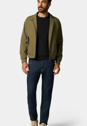 Olive green bomber jacket with a zip front, ribbed cuffs, and a black knit sweater beneath, paired with dark blue jeans and beige sneakers.