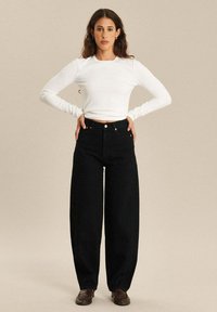 Zwarte wijdvallende jeans gecombineerd met een witte, nauwsluitende top met lange mouwen. De jeans hebben een hoge taille en een knoopsluiting aan de voorkant.