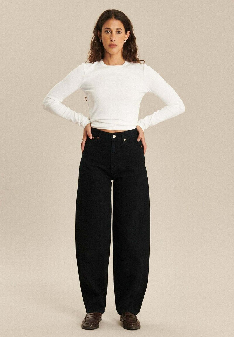 Zwarte wijdvallende jeans gecombineerd met een witte, nauwsluitende top met lange mouwen. De jeans hebben een hoge taille en een knoopsluiting aan de voorkant.