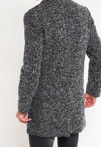Gray and black tweed coat with a textured pattern, long sleeves, standing collar, and rear vent. Styled with a relaxed fit and below-hip length.