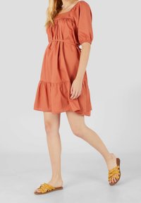 Robe en coton couleur rouille avec des manches bouffantes, taille cintrée et ourlet à volants. Associée à des sandales à lanières jaunes sur un fond clair.