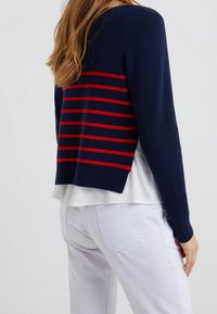 Navy knit sweater with red horizontal stripes, featuring long sleeves and a slightly cropped design. Layered over a white blouse.