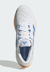 Zapatilla de running blanca y azul con parte superior de malla, cordones y suela naranja, vista desde arriba, texto "SUPERNOVA" visible en el interior.