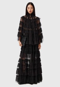 Schwarzes, gestuftes Maxikleid aus transparentem Stoff mit Rüschendetails, langen Ärmeln und hohem Halsausschnitt. Kombiniert mit einer schwarzen, strukturierten Clutch.