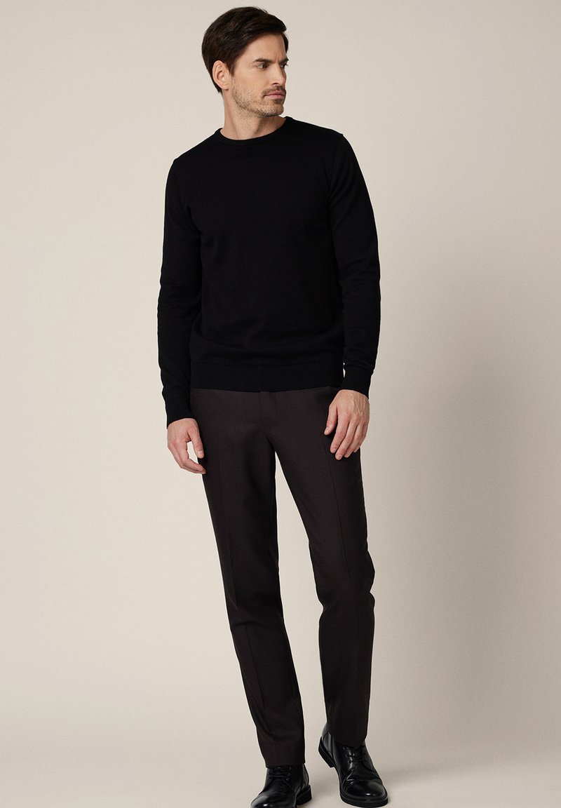 Czarny dzianinowy sweter z okrągłym dekoltem, zestawiony z dopasowanymi ciemnobrązowymi spodniami. Model stoi swobodnie, nosząc czarne formalne buty.