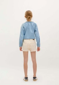 Blouse en denim bleu à manches longues avec un col froncé, associée à un short blanc effiloché et des chaussures en toile noires avec des semelles blanches.