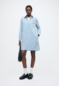 Rochie cu mâneci lungi de culoare albastru deschis, cu guler, țesătură texturată, buzunar la piept și buzunare laterale. Asortată cu pantofi loafer negri și șosete albe.