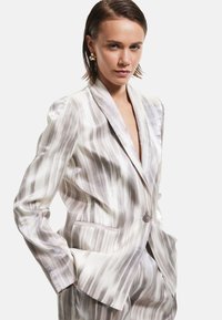 Blazer en tissu léger et soyeux avec un motif de rayures ondulées blanches et grises. Il possède un col à revers, une fermeture à bouton unique et des poches.